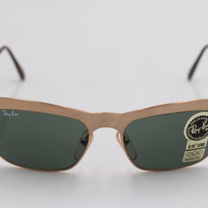 May include: Gold-toned Ray-Ban sunglasses with dark green lenses. The frames have a distinctive shape, and the lenses are slightly curved. A sticker on the lens reads "Ray-Ban" and "6-15 LENS". The sunglasses have a classic, retro style.