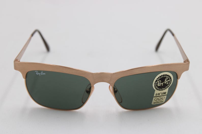 May include: Gold-toned Ray-Ban sunglasses with dark green lenses. The frames have a distinctive shape, and the lenses are slightly curved. A sticker on the lens reads "Ray-Ban" and "6-15 LENS". The sunglasses have a classic, retro style.