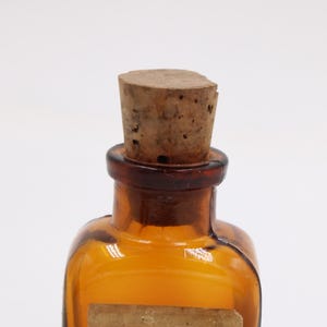 May include: Amber-colored glass bottle with a cork stopper. The bottle has a square shape with rounded edges and a dark brown neck. The cork is light brown and cylindrical. A small, rectangular label is affixed to the front of the bottle.