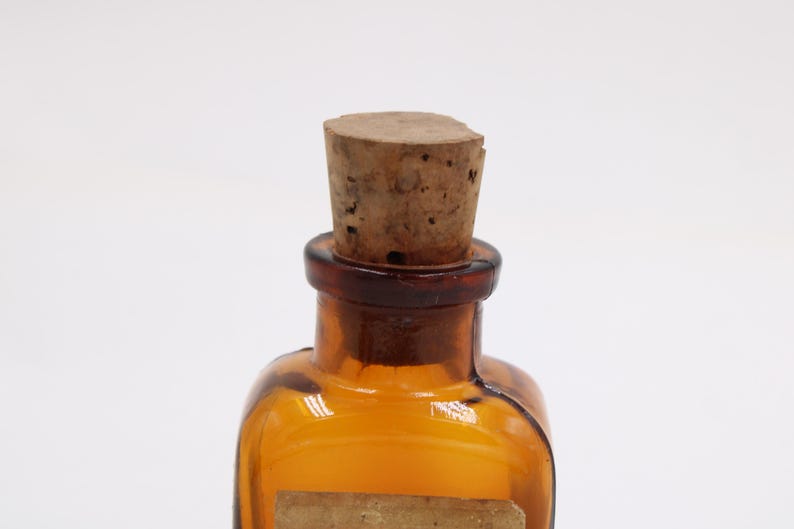May include: Amber-colored glass bottle with a cork stopper. The bottle has a square shape with rounded edges and a dark brown neck. The cork is light brown and cylindrical. A small, rectangular label is affixed to the front of the bottle.