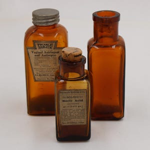 May include: Three antique amber glass medicine bottles. The bottles have labels with text, including "Vaginal Astringent and Antiseptic" and "Boric Acid". One bottle has a metal cap, and another has a cork stopper. The bottles are of varying sizes.