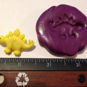 May include: A yellow plastic dinosaur figurine with a purple polymer clay mold of the same dinosaur. The mold is about 2 inches in diameter. The ruler in the background shows the size of the dinosaur.