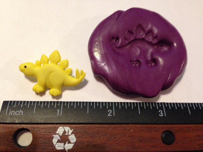 May include: A yellow plastic dinosaur figurine with a purple polymer clay mold of the same dinosaur. The mold is about 2 inches in diameter. The ruler in the background shows the size of the dinosaur.