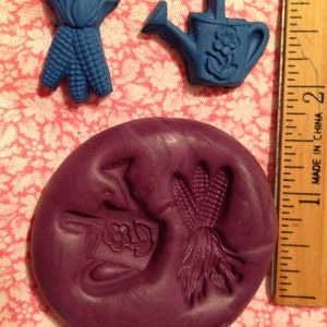May include: A purple silicone mold with two impressions: a watering can with a flower design and a bundle of corn. The mold is next to a ruler.