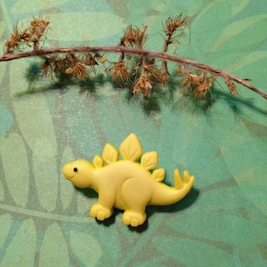 May include: A yellow plastic dinosaur toy with a spiky back and a small black eye. The dinosaur is on a green background with a brown twig.