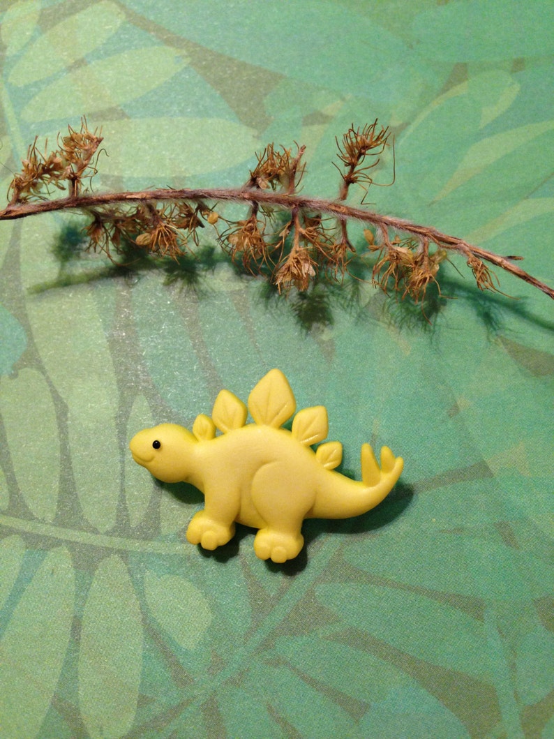 May include: A yellow plastic dinosaur toy with a spiky back and a small black eye. The dinosaur is on a green background with a brown twig.