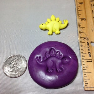 May include: A purple silicone mold with a raised design of a yellow dinosaur. The mold is about 2 inches in diameter. A yellow dinosaur figurine is shown for size comparison.