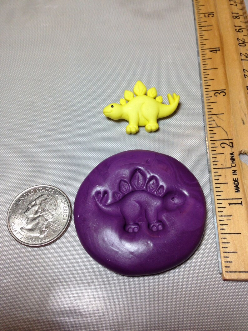 May include: A purple silicone mold with a raised design of a yellow dinosaur. The mold is about 2 inches in diameter. A yellow dinosaur figurine is shown for size comparison.