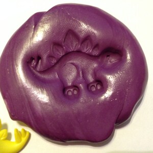 May include: Purple play dough with a dinosaur imprint. The dinosaur is facing right and has a spiky back.