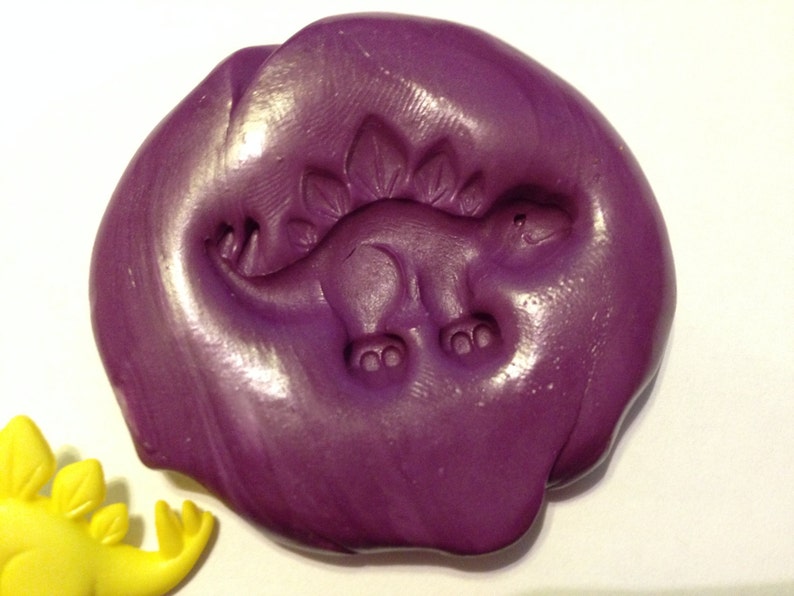 May include: Purple play dough with a dinosaur imprint. The dinosaur is facing right and has a spiky back.