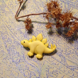 May include: A yellow plastic dinosaur toy with a spiky back and a friendly face. The dinosaur is sitting on a patterned background.