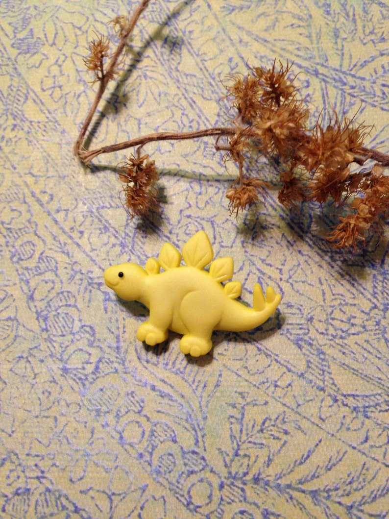 May include: A yellow plastic dinosaur toy with a spiky back and a friendly face. The dinosaur is sitting on a patterned background.