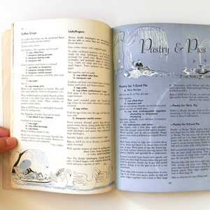 May include: An open vintage cookbook showing recipes for coffee crisps, ladyfingers, and pies. The pages have illustrations and text, with the title "Pastry & Pies". A hand is holding the book open.