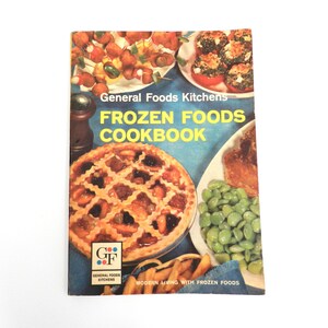 May include: A vintage General Foods Kitchens Frozen Foods Cookbook with a blue cover featuring colour photographs of various prepared dishes including a fruit pie, green beans, and appetizers, with yellow text reading Frozen Foods Cookbook.