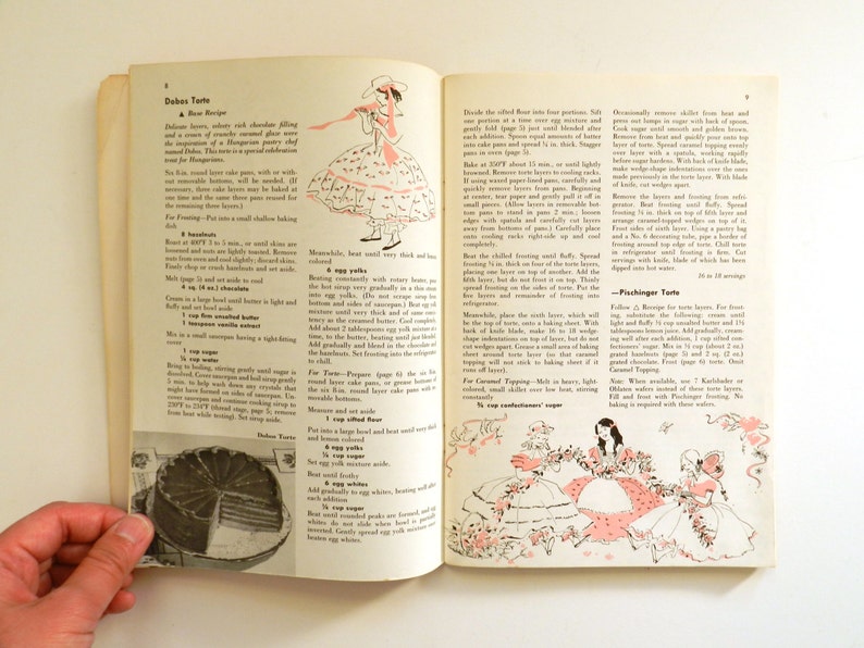 May include: A vintage cookbook open at a recipe for Dobos Torte, with illustrations of women in dresses. The pages are yellowed, and the illustrations are in pink and black. A hand holds the book open, showing the recipe.