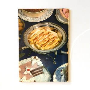May include: A vintage cookbook cover showing an assortment of desserts on a dark blue tablecloth. Rolled crepes are on a silver platter. Cakes and tarts are also visible. The image has a retro style.