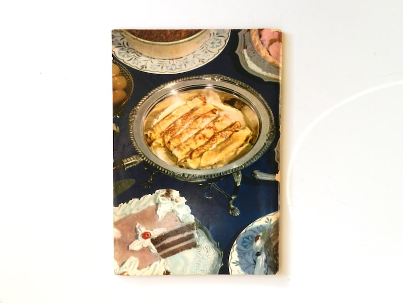 May include: A vintage cookbook cover showing an assortment of desserts on a dark blue tablecloth. Rolled crepes are on a silver platter. Cakes and tarts are also visible. The image has a retro style.