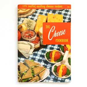 May include: A vintage cookbook titled "The Cheese Cookbook" with an orange banner. The cover features a collage of cheese varieties, sandwiches, and tomato dishes. The background has a blue and white checkered pattern.