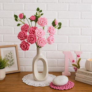 May include: A display of pink and light pink crocheted roses in a white vase. The arrangement is on a wooden table with a framed botanical print, a small potted plant, and decorative items. The roses have green leaves.