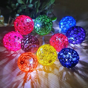 Könnte beinhalten: Eine Lichterkette mit bunten, gehäkelten Kugeln. Die Kugeln sind in den Farben Rot, Grün, Blau, Lila, Orange und Gelb gehalten. Die Lichter sind mit einem durchsichtigen Draht verbunden.