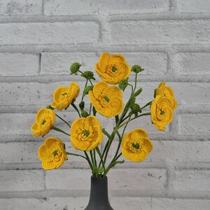 May include: A display of yellow crochet flowers with green stems and buds, presented in a black vase. The handmade flowers feature intricate petal details, set against a white brick wall.