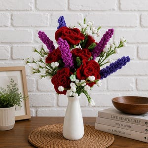 Puede incluir: Un ramo artesanal de flores de ganchillo en un jarrón blanco. El arreglo presenta vibrantes rosas rojas, lavanda morada y azul, y delicado aliento de bebé blanco. El jarrón está sobre una estera tejida marrón, con libros y un cuenco de madera en el fondo.