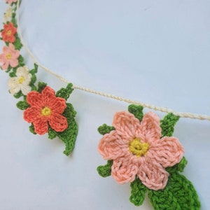 May include: Crocheted floral garland featuring peach, coral, and white flowers with yellow centers and green leaves. The flowers are attached to a cream-colored string. This handmade garland is perfect for home decor or special events.