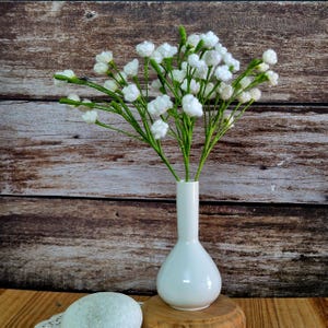 May include: A white ceramic vase holds a bouquet of white baby's breath flowers with green stems. The vase has a rounded base and a long, slender neck. The arrangement sits on a wooden surface against a wood-paneled backdrop.