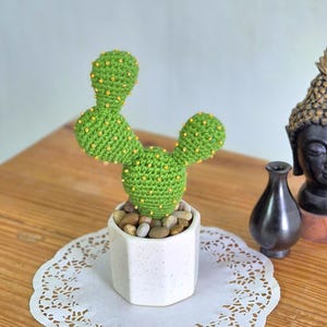 以下が含まれることがあります： 黄色いビーズのアクセントが付いた手作りの緑色の鉤針編みのサボテンが、小さな石で満たされた白い六角形のポットに入っています。 サボテンは白いドイリーの上にあり、木製の表面に置かれています。 背景には装飾品があります。