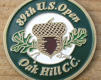 Op de afbeelding: Een groene en gouden golftoernooi-herinneringsmunt met de tekst "89th U.S. Open Oak Hill C.C." en een eikelontwerp.