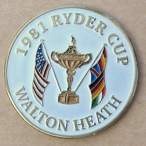 May include: A white, round, metal coin with gold lettering and a gold-colored trophy. The coin reads "1981 Ryder Cup Walton Heath". The trophy is flanked by two flags, one American and one with the colors of the United Kingdom.