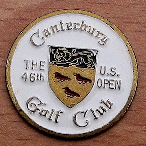 Op de afbeelding: Een witte en gouden golfbalmarker met de tekst "Canterbury Golf Club" en "The 46th U.S. Open" rond een wapenschild met een leeuw en vogels.