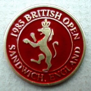 Op de afbeelding: Een ronde rode en gouden pin met de tekst "1985 British Open Sandwich, England" en een gouden leeuw met een kroon op zijn kop.