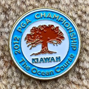 Op de afbeelding: Een ronde blauwe en gouden pin met de tekst "2012 PGA Championship The Ocean Course Kiawah" en een bruine boomillustratie.