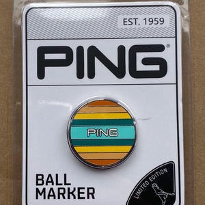 May include: A PING ball marker in a clear plastic package. The marker is round with a striped design in shades of orange, yellow, and teal. The word "PING" is printed on the marker and the packaging. The packaging also says "EST. 1959" and "LIMITED EDITION".