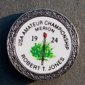 Könnte beinhalten: Ein silberner Golfball-Marker mit weißem Hintergrund und schwarzem Text. Der Text lautet "USA Amateur Championship Merion 19 24 Robert T. Jones". Ein grüner Baum und ein rot-weißer Golfschläger befinden sich in der Mitte des Markers.