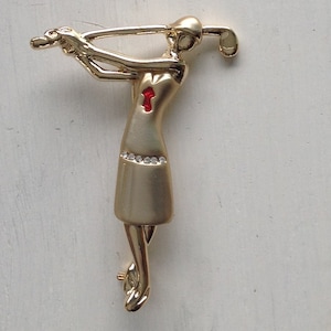 Könnte beinhalten: Goldfarbene Metallbrosche zum Thema Golf. Die Brosche zeigt einen Golfer im Schwung, mit einem roten Akzent auf dem Kleid und einer Reihe kleiner Strasssteine. Der Golfer hält einen Golfschläger. Ein stilvolles Accessoire für Golfbegeisterte.