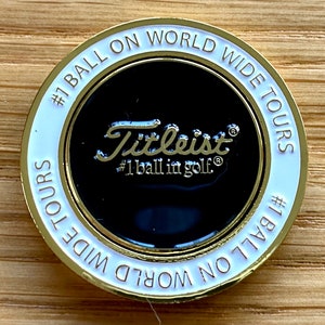 Könnte beinhalten: Ein goldfarbener Golfball-Marker mit weißem Rand. Der Marker zeigt den Text "#1 BALL ON WORLD WIDE TOURS" und das Titleist-Logo mit dem Text "#1 ball in golf®".