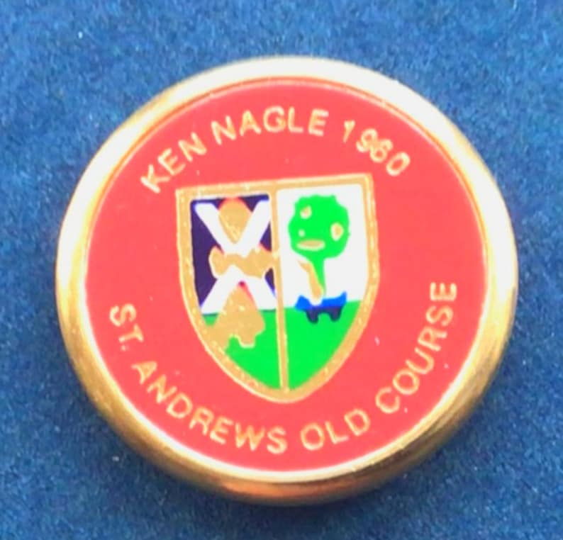 K&ouml;nnte beinhalten: Eine runde rote und goldene Anstecknadel mit dem Text "Ken Nagle 1980 St Andrews Old Course" und einem bunten Wappen in der Mitte.