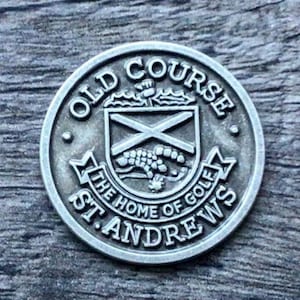 Könnte beinhalten: Eine silberfarbene runde Münze mit den Worten "Old Course" und "St. Andrews" am Rand. In der Mitte befindet sich ein Wappen mit einem Kreuz und einem kleinen Tier.