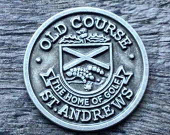 Op de afbeelding: Een zilverkleurige ronde munt met de woorden "Old Course" en "St. Andrews" rond de rand. Het midden heeft een wapenschild met een kruis en een klein dier.