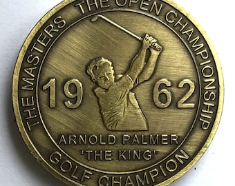 May include: A gold-colored coin commemorating Arnold Palmer's 1962 win at The Masters. The coin features a relief image of Palmer swinging a golf club, with the text "THE MASTERS THE OPEN CHAMPIONSHIP 19 62 ARNOLD PALMER 'THE KING' GOLF CHAMPION" around the edge.