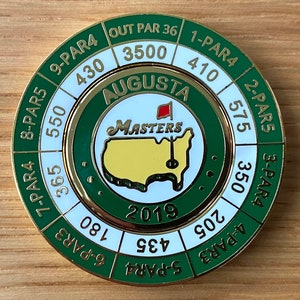 May include: A gold-colored golf ball marker with the Augusta Masters logo, a map of the United States in yellow, and the year 2019. The marker has a green and white ring with hole yardage and par information.