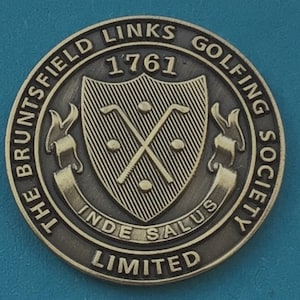 Könnte beinhalten: Eine goldfarbene Metallmünze mit dem Text "The Bruntsfield Links Golfing Society Limited" und "1761" am Rand. Die Mitte zeigt ein Schild mit gekreuzten Golfschlägern und dem Text "Inde Salus".