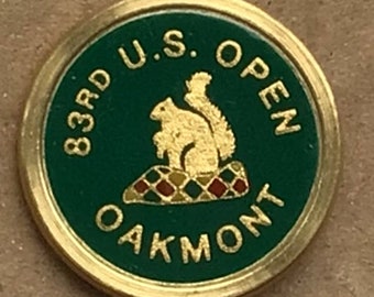 以下が含まれることがあります： 緑色の背景に金色の丸いピン。ピンには「83rd U.S. OPEN OAKMONT」という文字と、チェック柄の上に座っている金色のリスが描かれている。