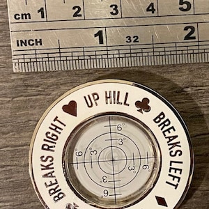 Puede incluir: Un nivel circular blanco con un borde negro y texto negro. El texto dice "Up Hill Breaks Left Down Hill Breaks Right". El nivel tiene un nivel de burbuja circular con marcas para 3, 6 y 9 grados.