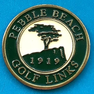 Peut inclure: Un marqueur de balle de golf rond avec un design vert et doré. Le marqueur porte l'inscription "PEBBLE BEACH GOLF LINKS" sur le bord, avec "1919" au centre. Un arbre et un graphique de falaise sont au centre.