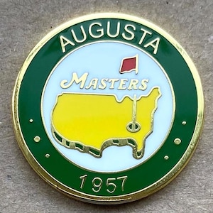 May include: A round, gold-rimmed pin with a green border featuring the text "AUGUSTA" at the top. The center has a white background with "Masters" in script, a red flag, and a yellow map of the U.S. with a golf pin. The year "1957" is at the bottom.