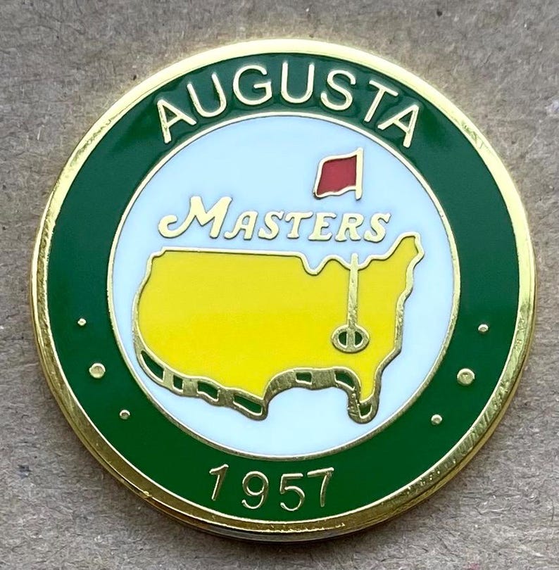 May include: A round, gold-rimmed pin with a green border featuring the text "AUGUSTA" at the top. The center has a white background with "Masters" in script, a red flag, and a yellow map of the U.S. with a golf pin. The year "1957" is at the bottom.