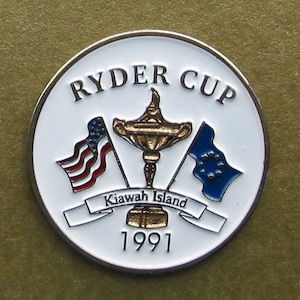 以下が含まれることがあります： 白い背景と金色の縁取りが施された、丸いライダーカップ記念コイン。コインには、ライダーカップのトロフィー、アメリカとヨーロッパの旗、そして「Ryder Cup」、「Kawah Island」、「1991」の文字が描かれています。
